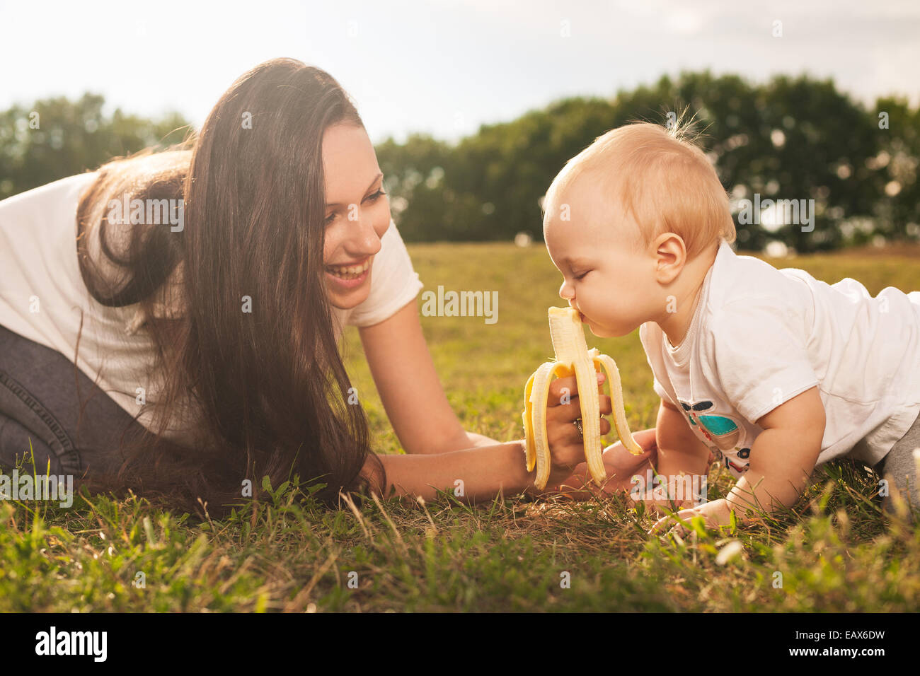 Young beautiful mother feeding her baby banana outdoors in sunlight Stock Photo