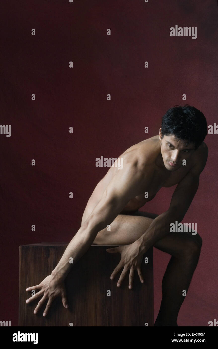 Nude man sitting, bending over to touch side of wooden pedestal Stock Photo