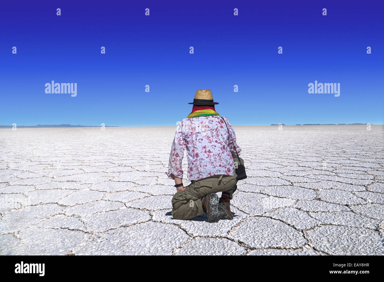 Traveler with straw hat and scarf in bolivian national colors kneels at the desert of Uyuni, Bolivia and looks at the wideness o Stock Photo