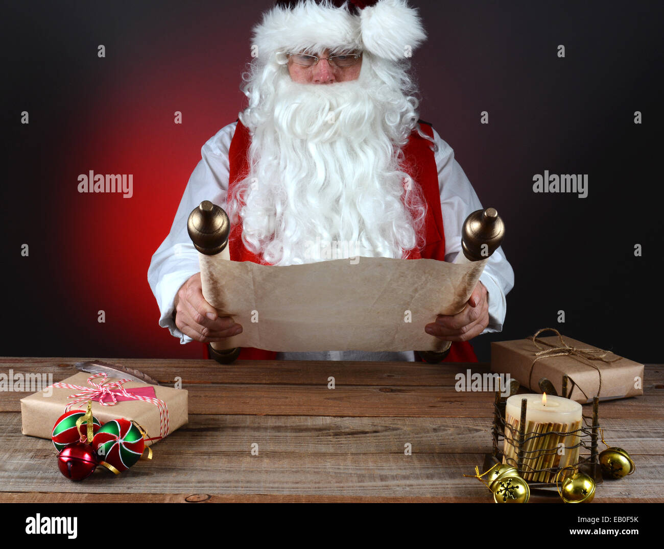 Santa Claus holding his Naughty and Nice list on a scroll of parchment paper. Santa is seated at a wood table with presents and Stock Photo
