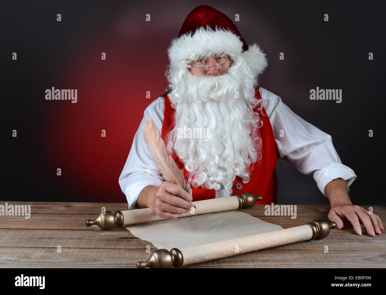 Santa Claus making his naughty and nice list on a scroll of parchment paper. Santa is seated at a rustic wood table. Horizontal Stock Photo