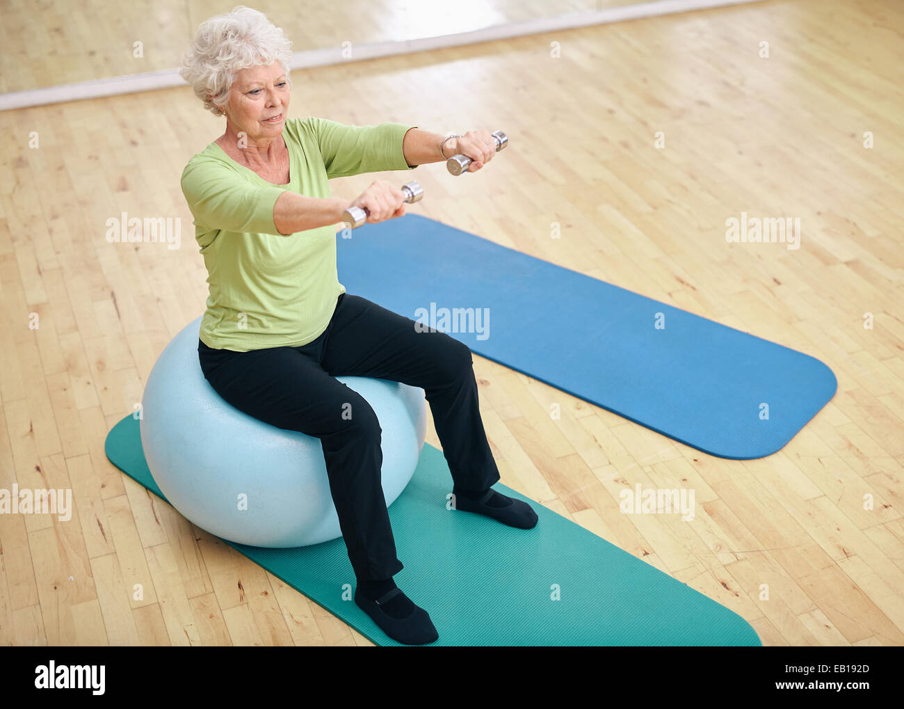 Senior female sitting on a fitness ball and lifting dumbbells. Old woman exercising with weights at gym. Stock Photo