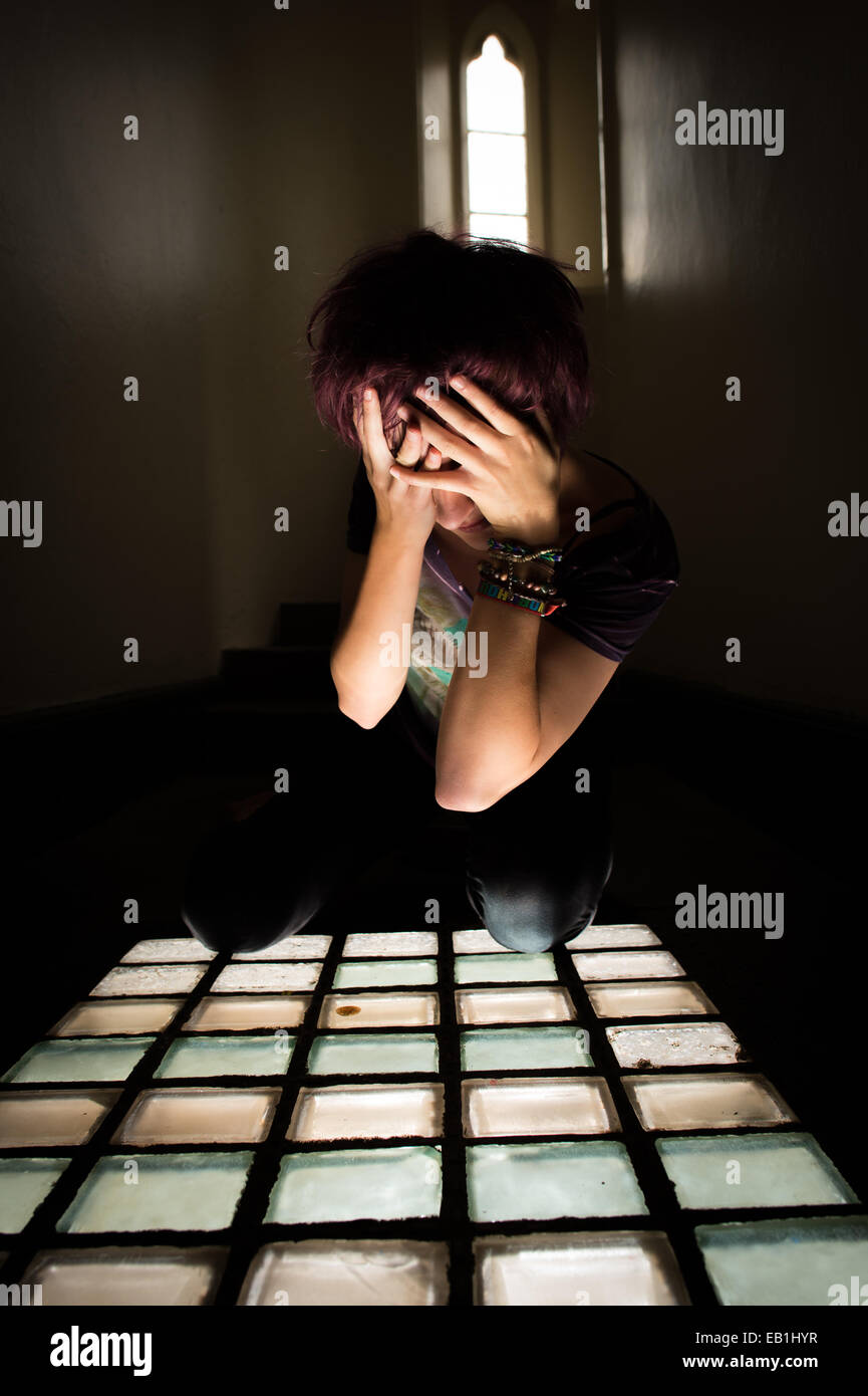 A despairing young teenage girl woman in a prison jail cell type location with light streaming through a barred window Stock Photo