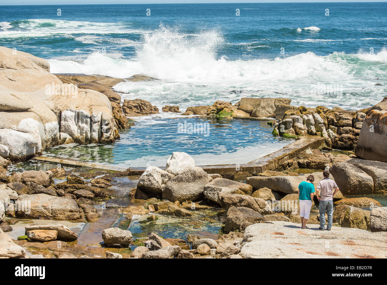 Two people standing on the rocks at Sea Point, Cape Town, near a man made tidal pool, watching the waves crashing in to the rock Stock Photo