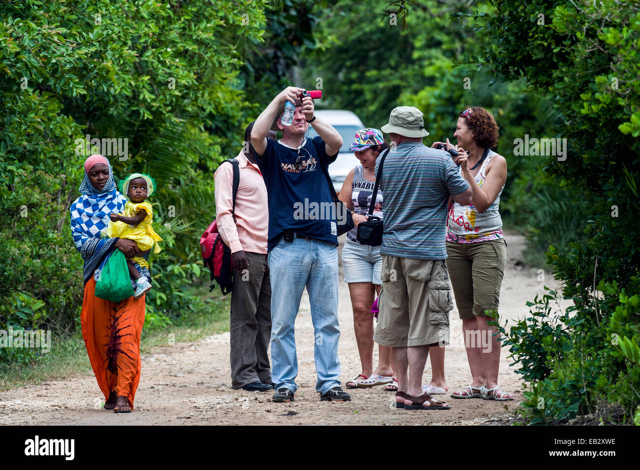 A villager and baby walk by tourists watching Zanzibar Red Colobus in a ...