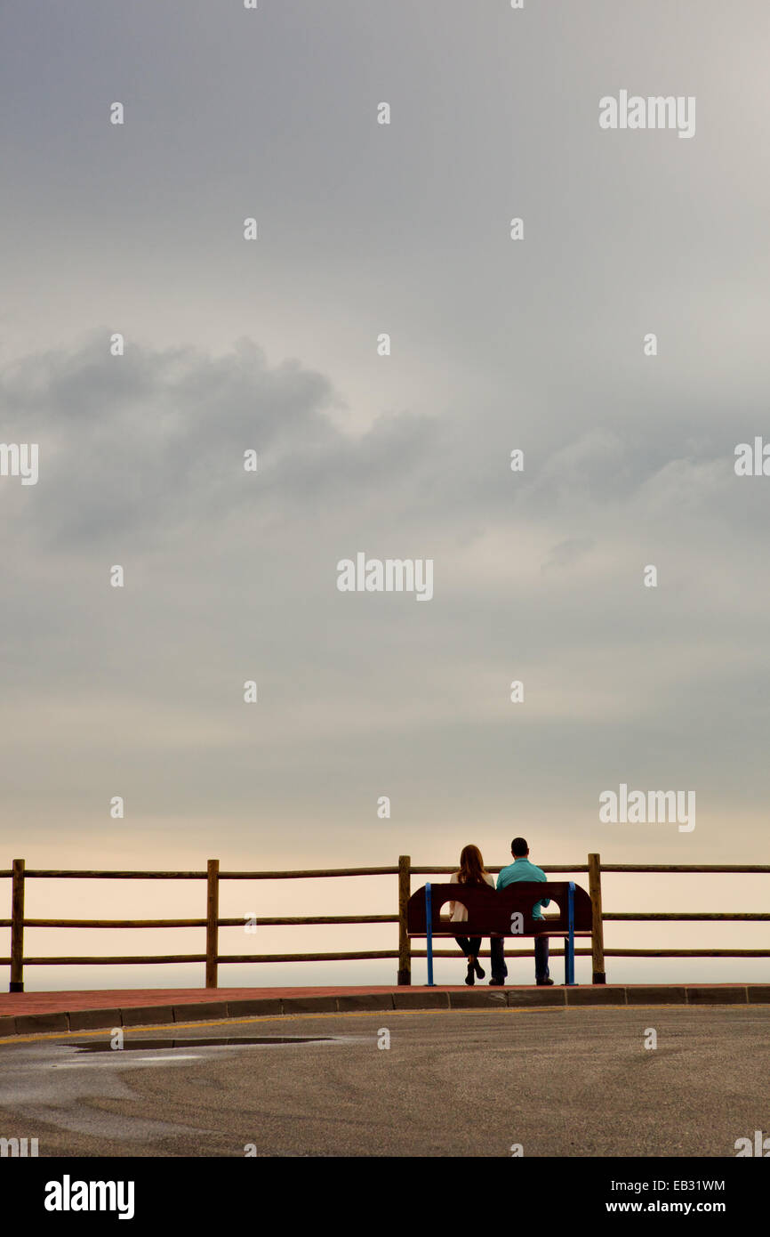 Silhouette of a heterosexual young couple on a calm and peaceful cloudy day, relaxing in front of the ocean view Stock Photo