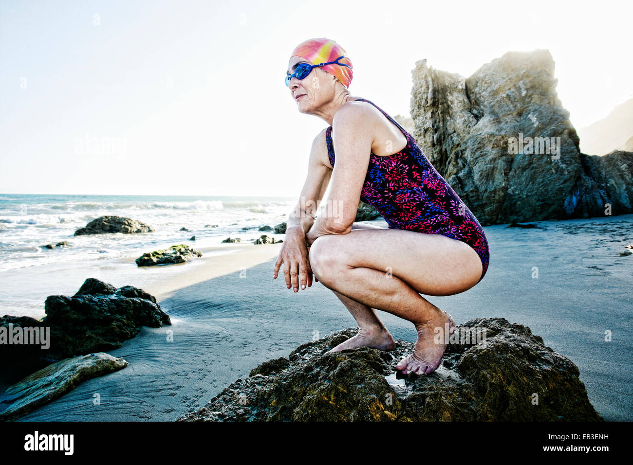 Older Caucasian woman crouching on rock on beach Stock Photo