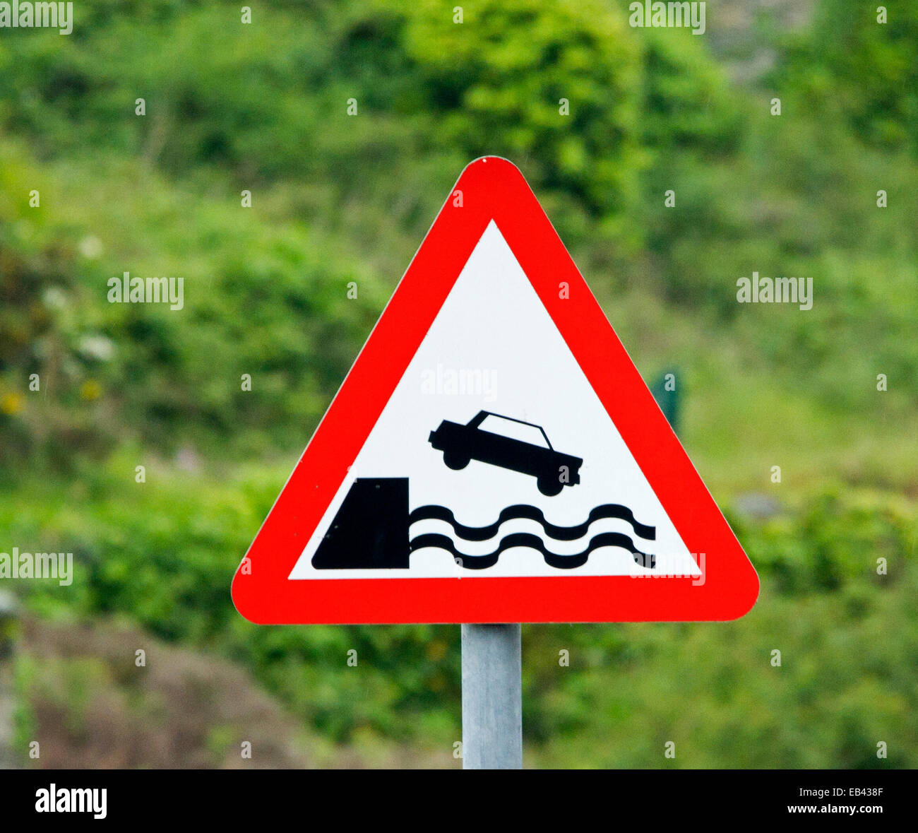 Humourous red edged triangular sign by harbour warning of danger of car falling over wall into ocean, on green background Stock Photo