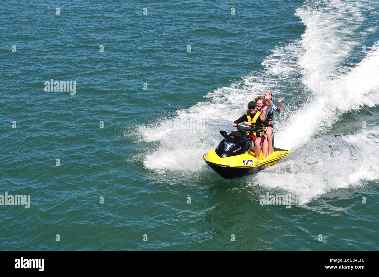 Family enjoying fast ride on jet sky creating significant wake wave on water surface. Stock Photo