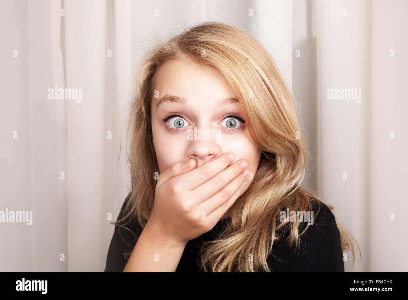 Beautiful blond Caucasian surprised girl opened her eyes wide and covers her mouth with her hands, closeup studio portrait Stock Photo