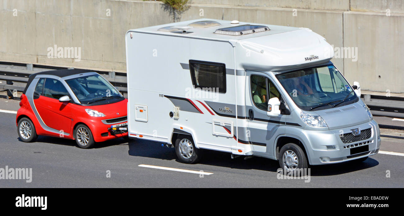 Side & front view of RV camper van motorhome recreational vehicle towing a red Smart Fortwo car driving along UK motorway Stock Photo
