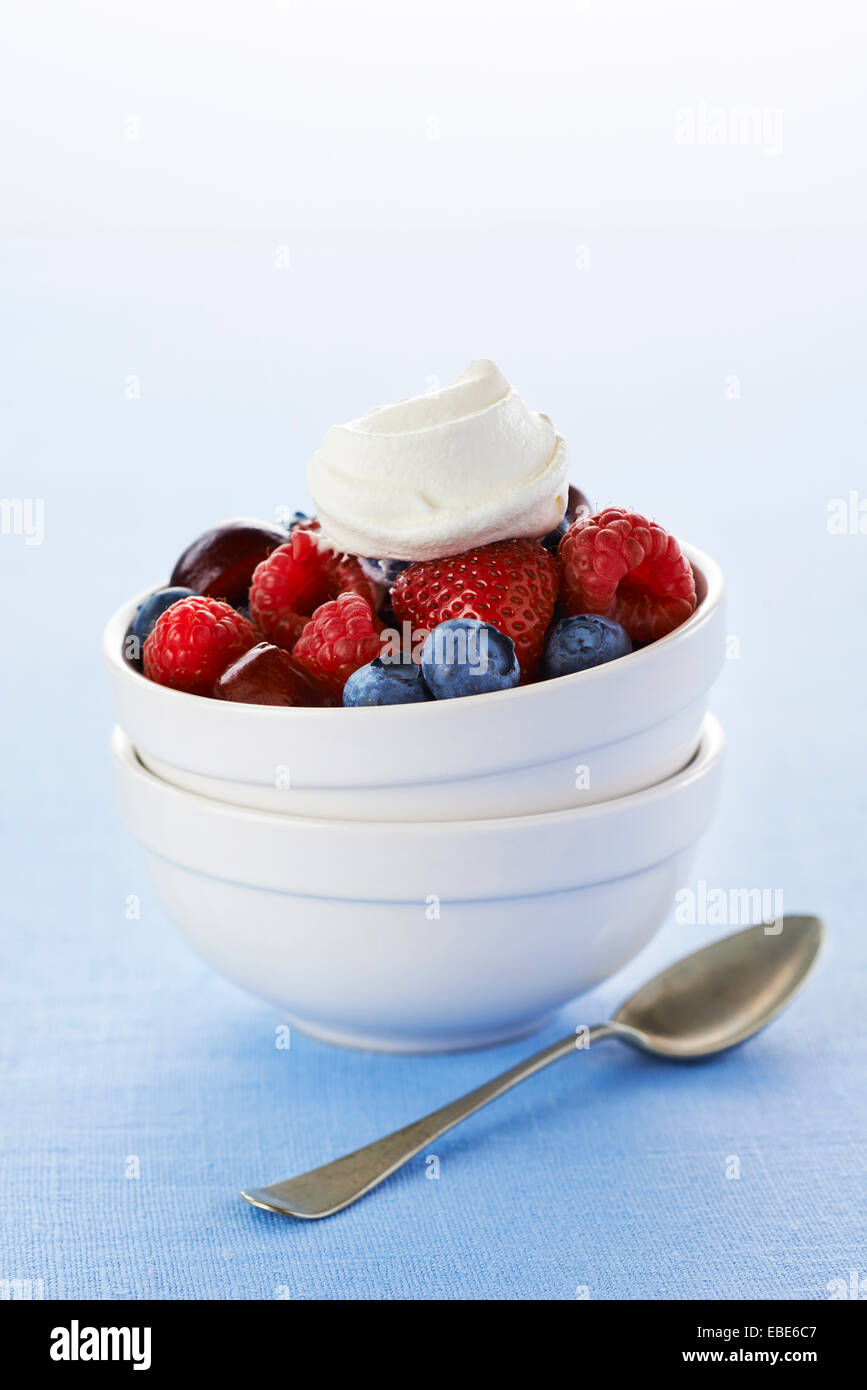 Bowl of Strawberries, Blueberries, Raspberries and Cherries with Whipped Cream and Spoon on Blue Background, Studio Shot Stock Photo