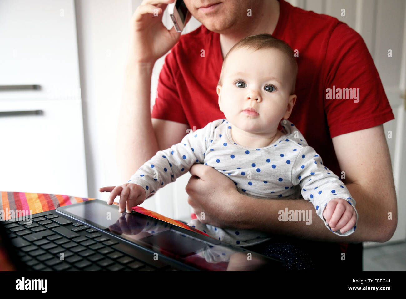 A father works on his laptop from home while holding his eight month old baby. Stock Photo
