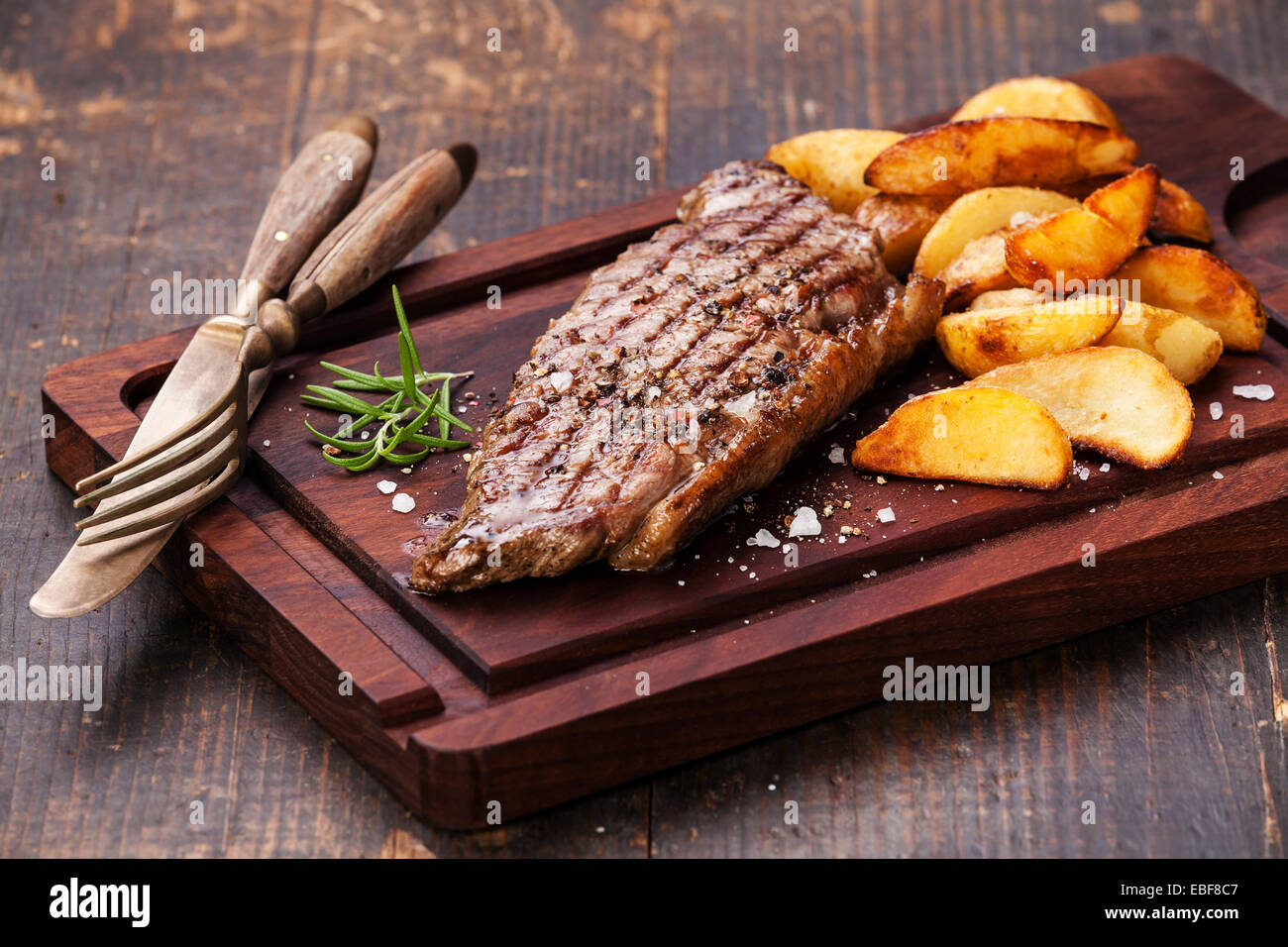 Well done grilled New York steak with roasted potato wedges on cutting board on dark wooden background Stock Photo