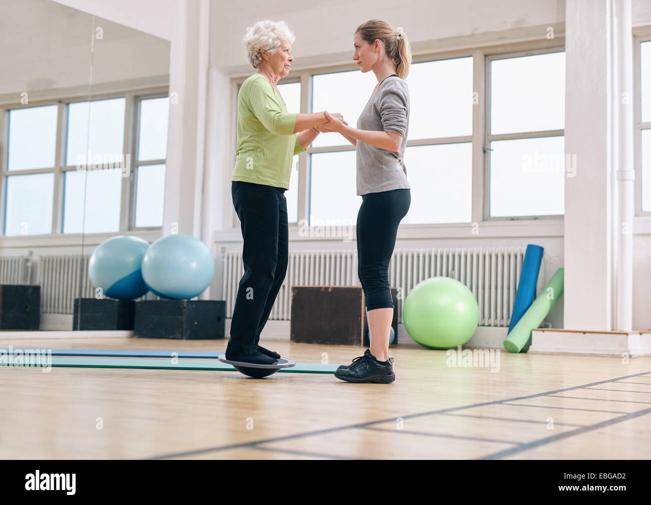 Female trainer helping senior woman standing on a balance board at gym