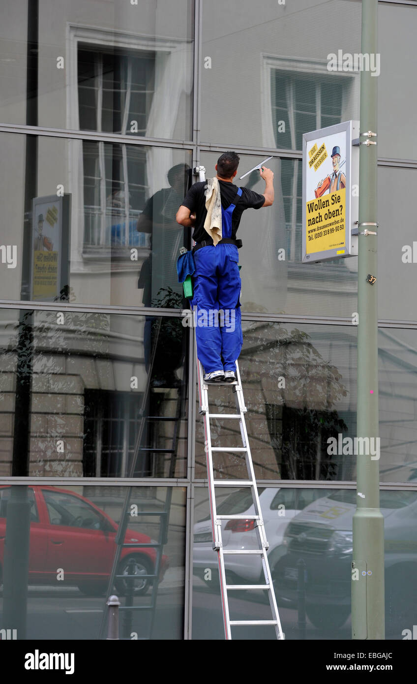 Window cleaner standing on a ladder cleaning a glass façade, next to advertising sign "Wollen Sie nach oben?", German for "do Stock Photo