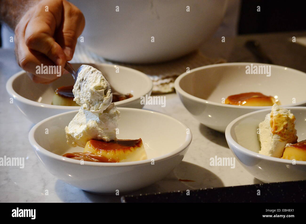 A chef's hand pouring cream to four servings of the typical Argentine dessert: flan with cream and caramel (dulce de leche) Stock Photo