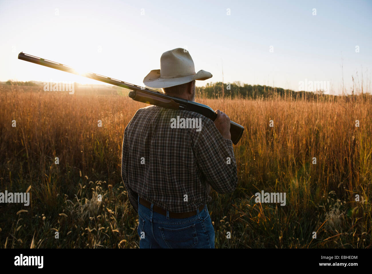 Senior male farmer with shotgun on shoulder in remote field at dusk