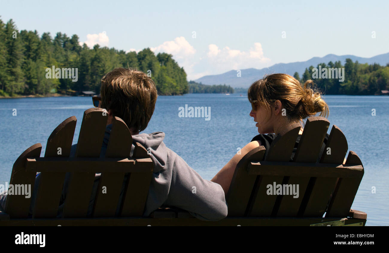 Young couple man woman sitting in double Adirondack chair over looking Long Lake New York USA. Adirondack State Park Adirondacks Stock Photo