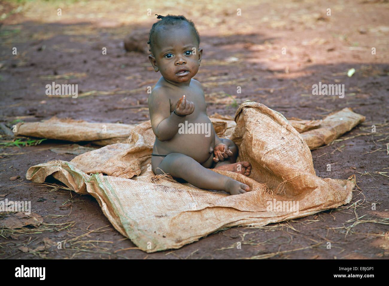 Female young child in a poor area sitting on a plastic bag with a belly that is bloated by worms and malnutrition, Uganda, Jinja Stock Photo