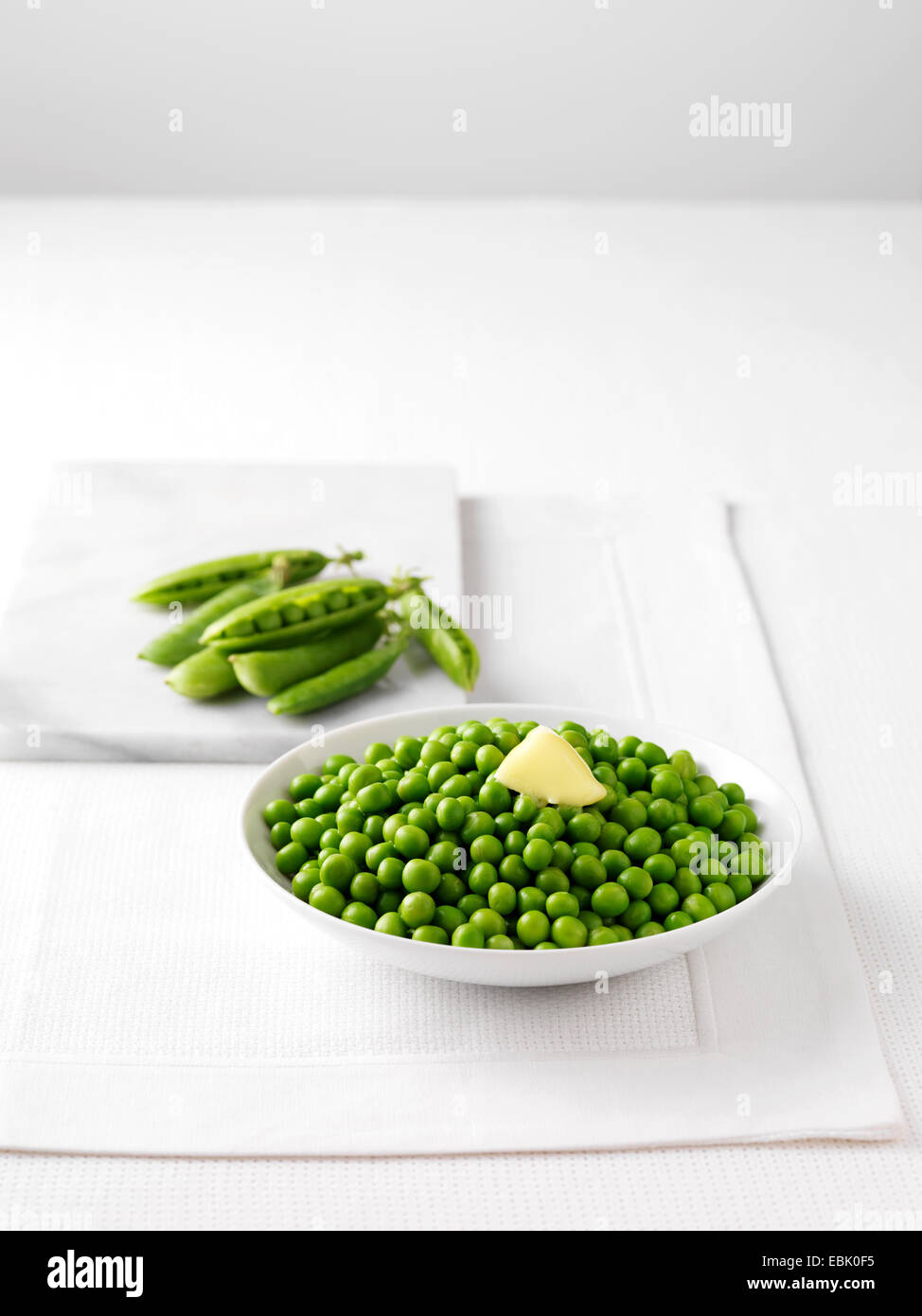 Raw pea pods on marble cutting board and bowl of boiled peas with butter Stock Photo