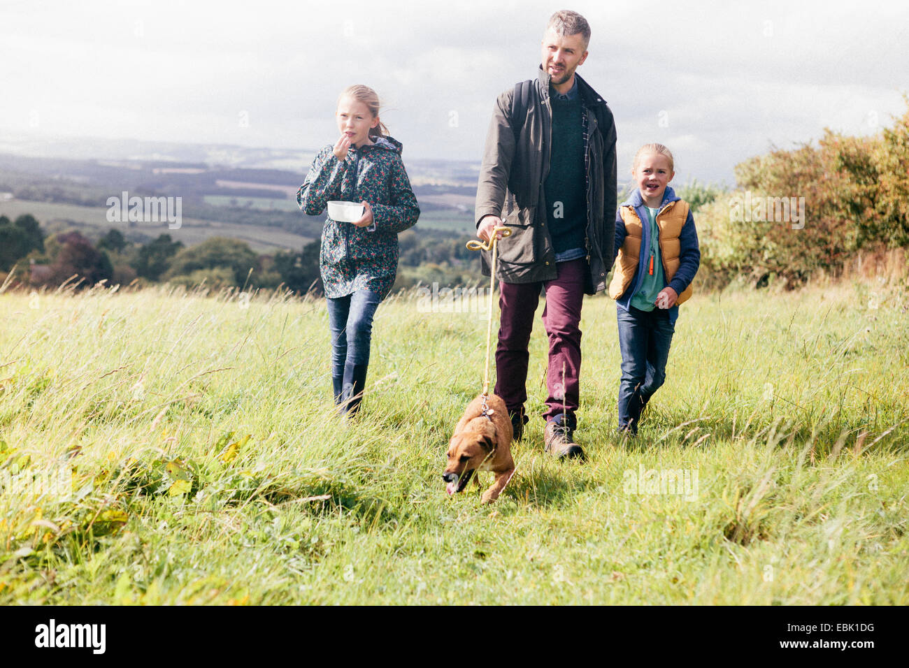 Father and two girls walking dog in field Stock Photo