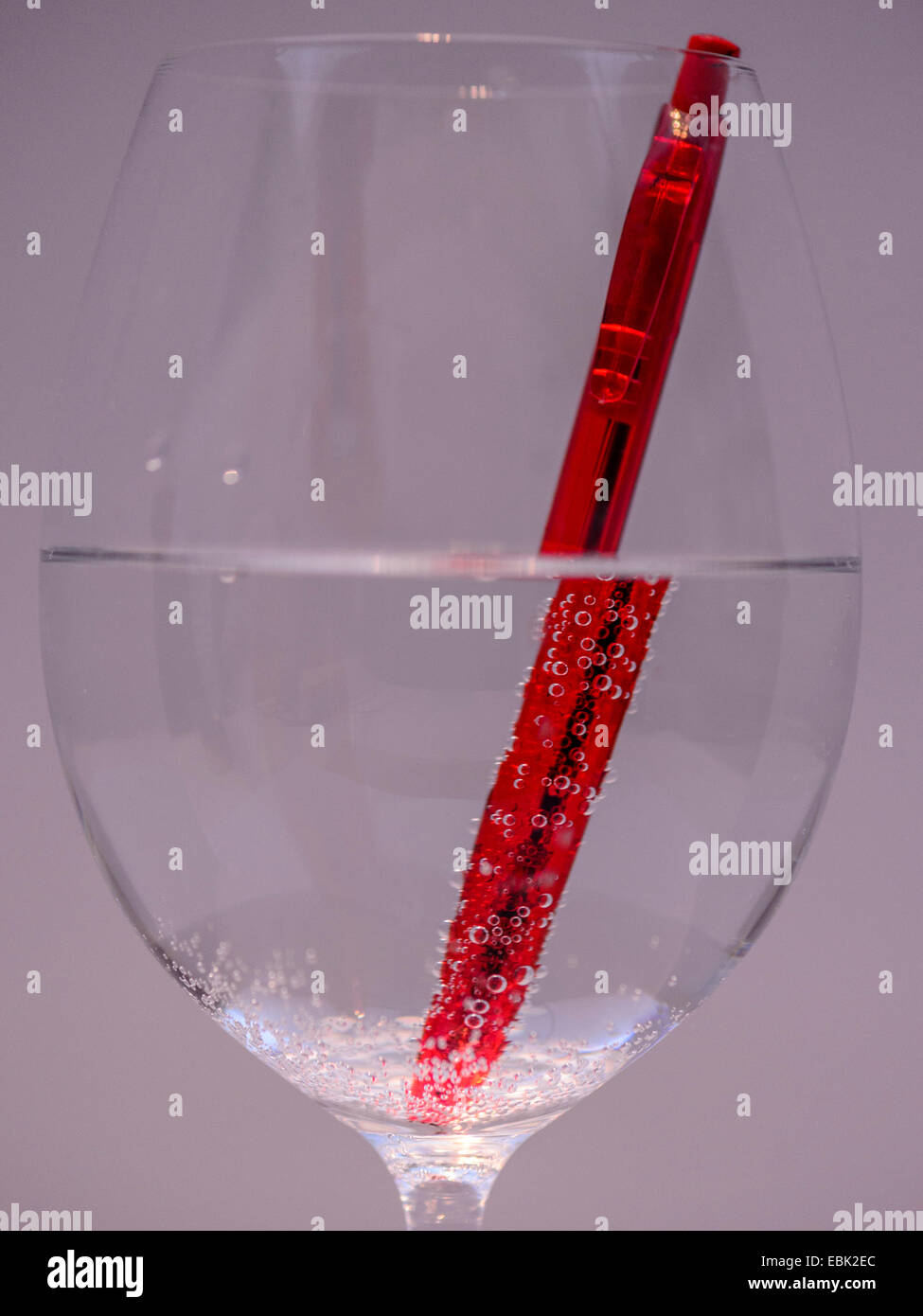 Studio macro image, vibrant red pen half immersed in a wine glass of sparkling water showing diffraction of the pen in water. Stock Photo