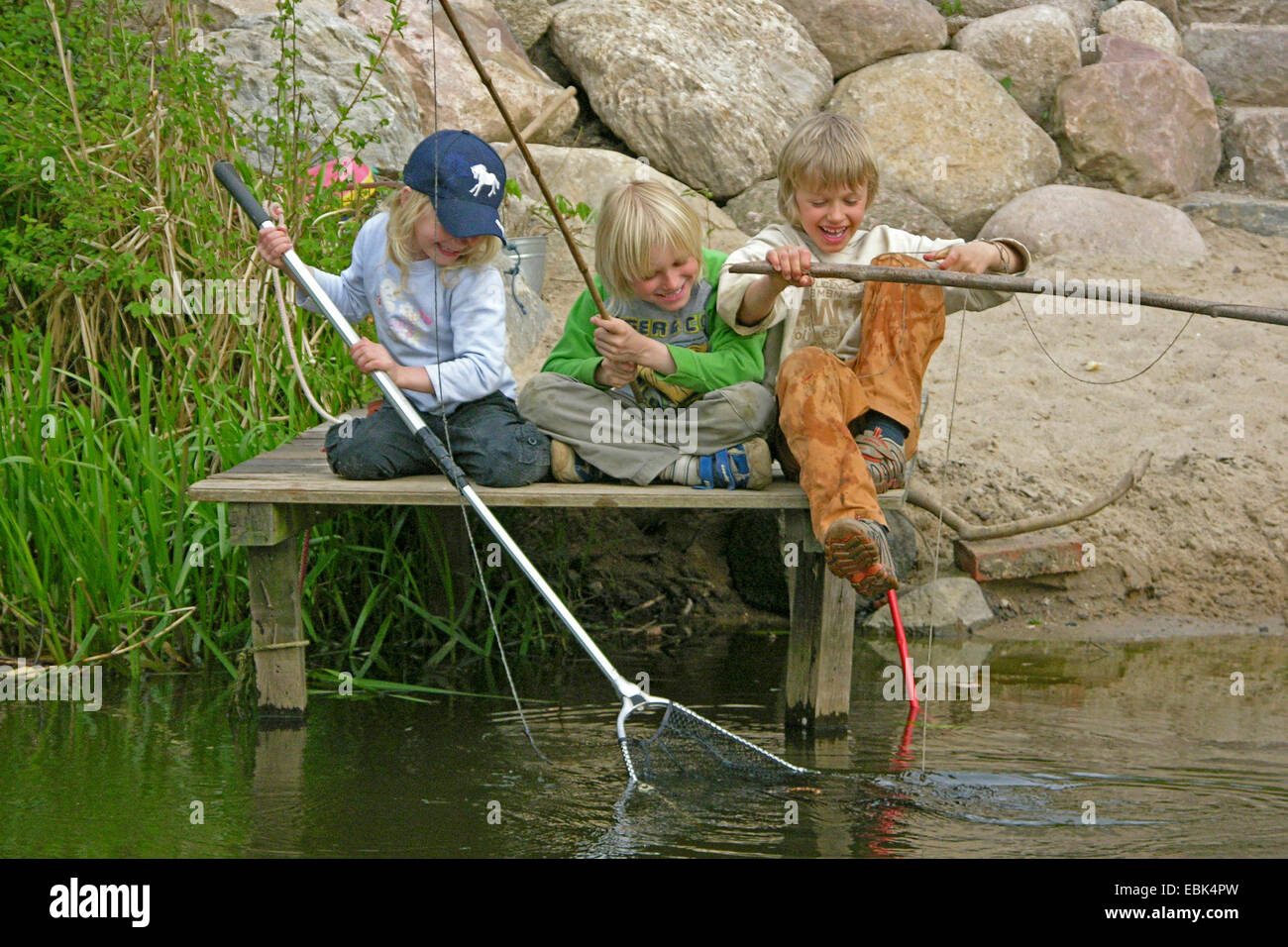 three children sitting on boardwalk, scooping and fishing, Germany Stock Photo