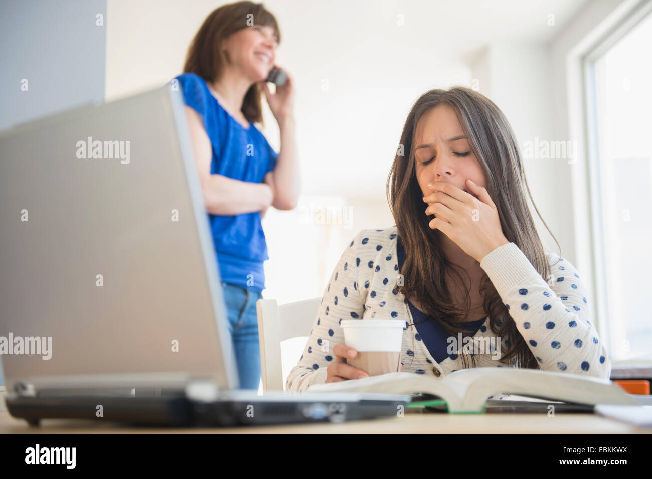 Tired teenage girl (14-15) yawning in front of textbook, mother on phone in background Stock Photo