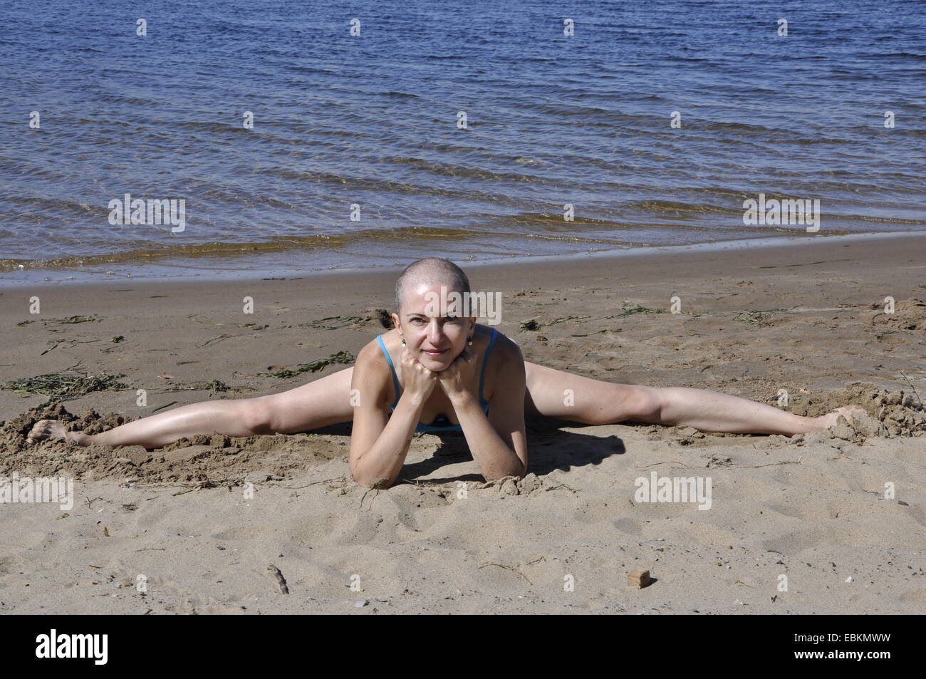 Bald woman sitting in split leaning on her elbows on a sandy river beach, smiling Stock Photo