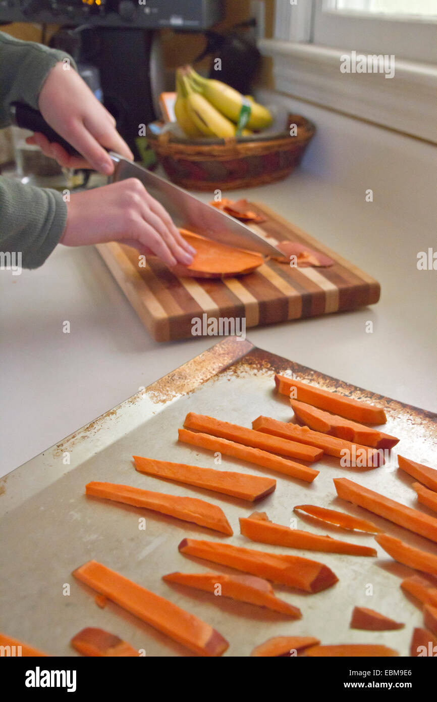 Young woman's hands cut sweet potato fries on a cutting board with uncooked fries on a baking sheet in the foreground Stock Photo