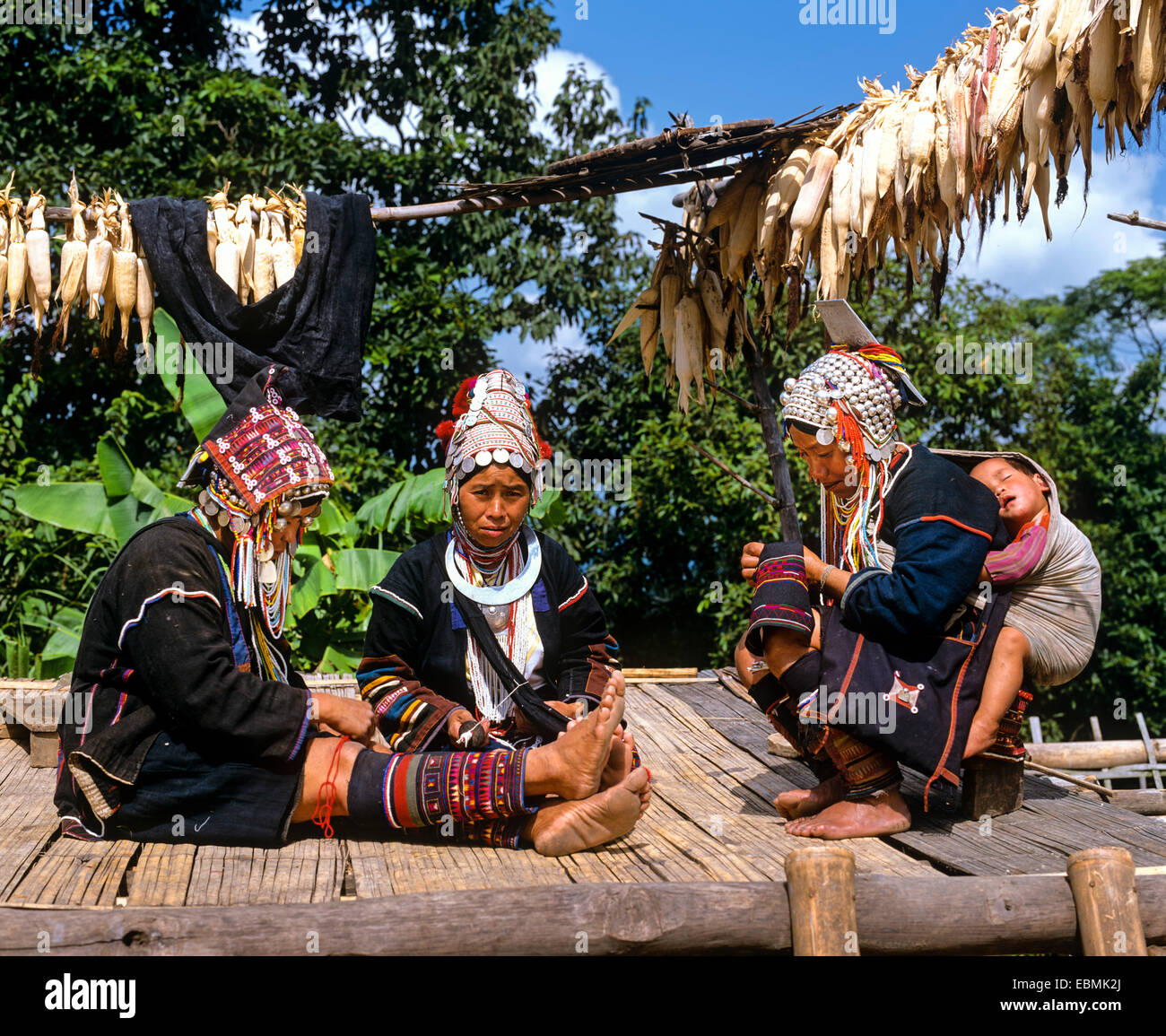 Akha women in a mountain village, in traditional costume and headdresses with silver coins, one carrying a sleeping child in a Stock Photo