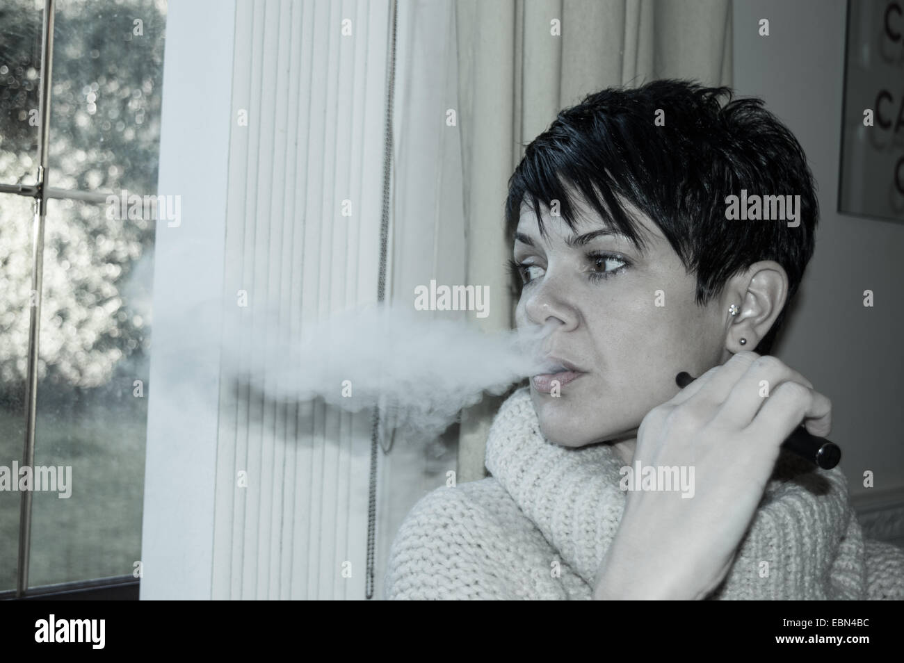 Beautiful short, dark haired woman vaping and looking out of a window deep in thought Stock Photo
