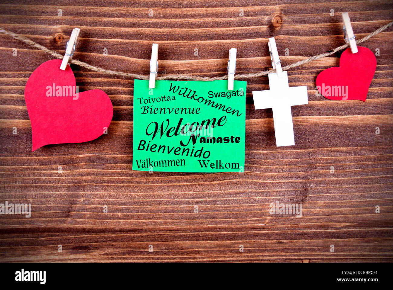 Green Tag Or Label With Hearts And Cross On A Line With Welcome In Different Or Many Languages On Wooden Background, Three Symbo Stock Photo