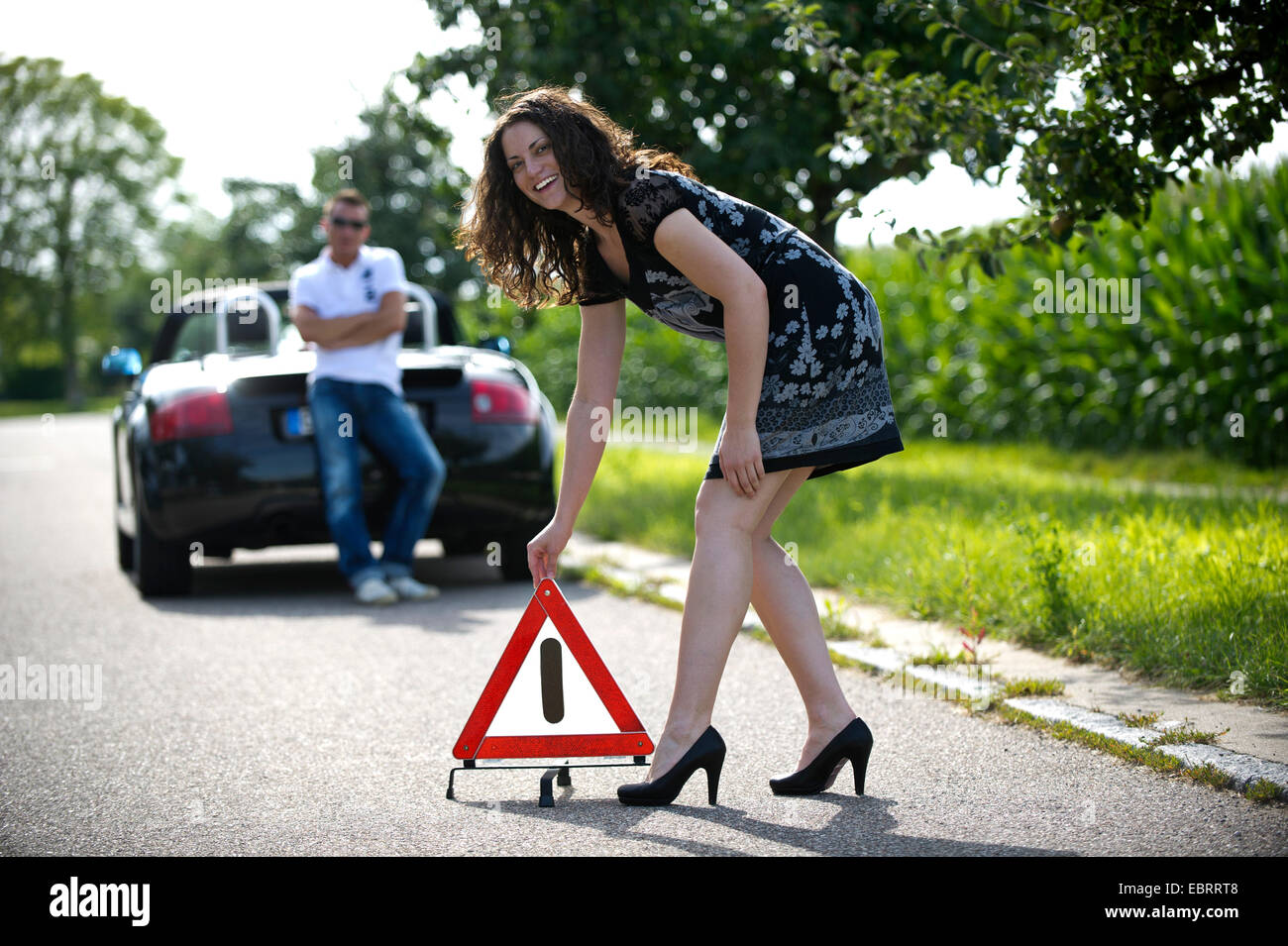 young woman putting up a warning triangle with a smile after a breakdown on a country road while taking it humorously, Germany Stock Photo