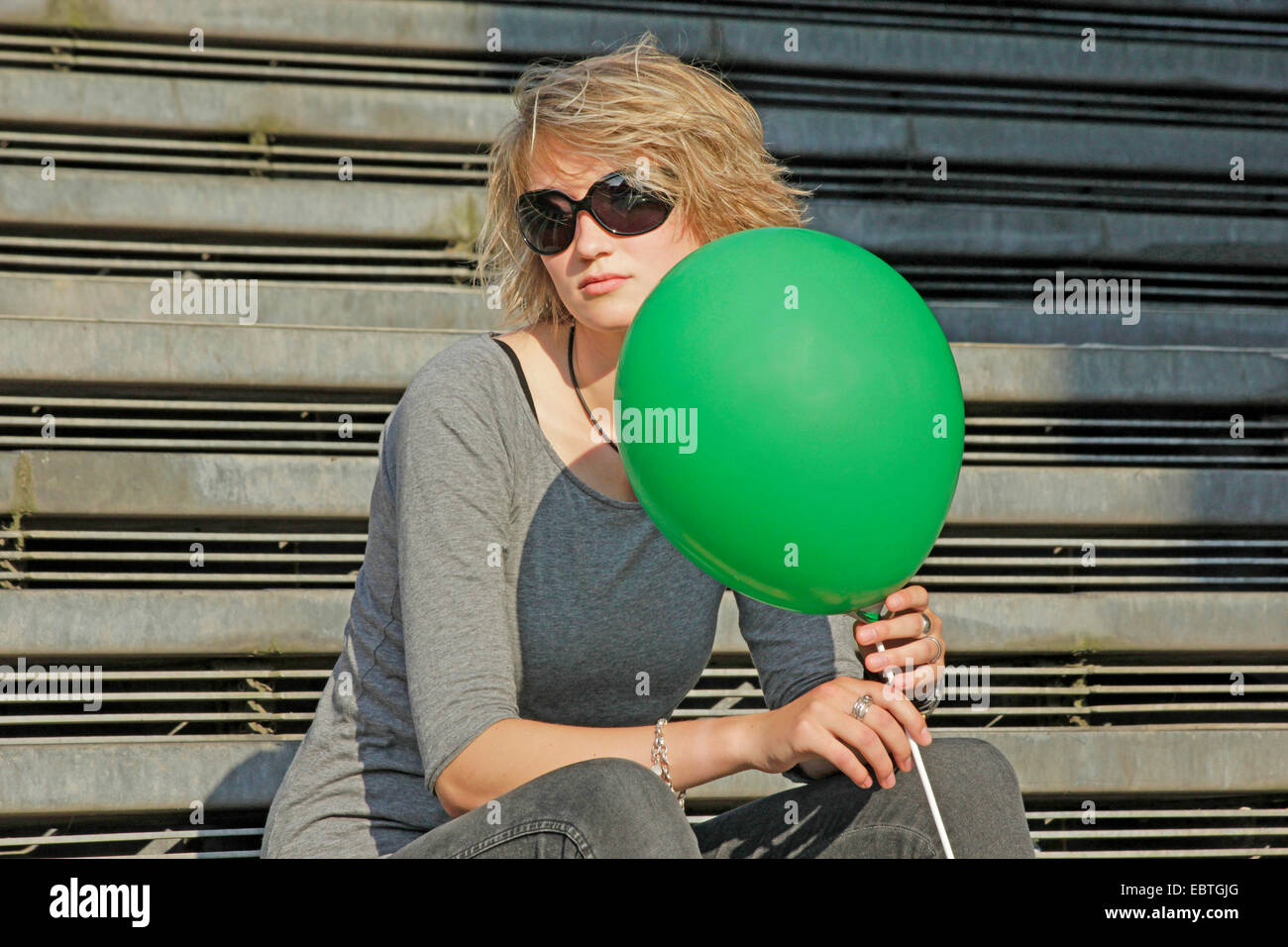 young blond woman with sunglasses and green balloon sitting on stairs, Germany Stock Photo