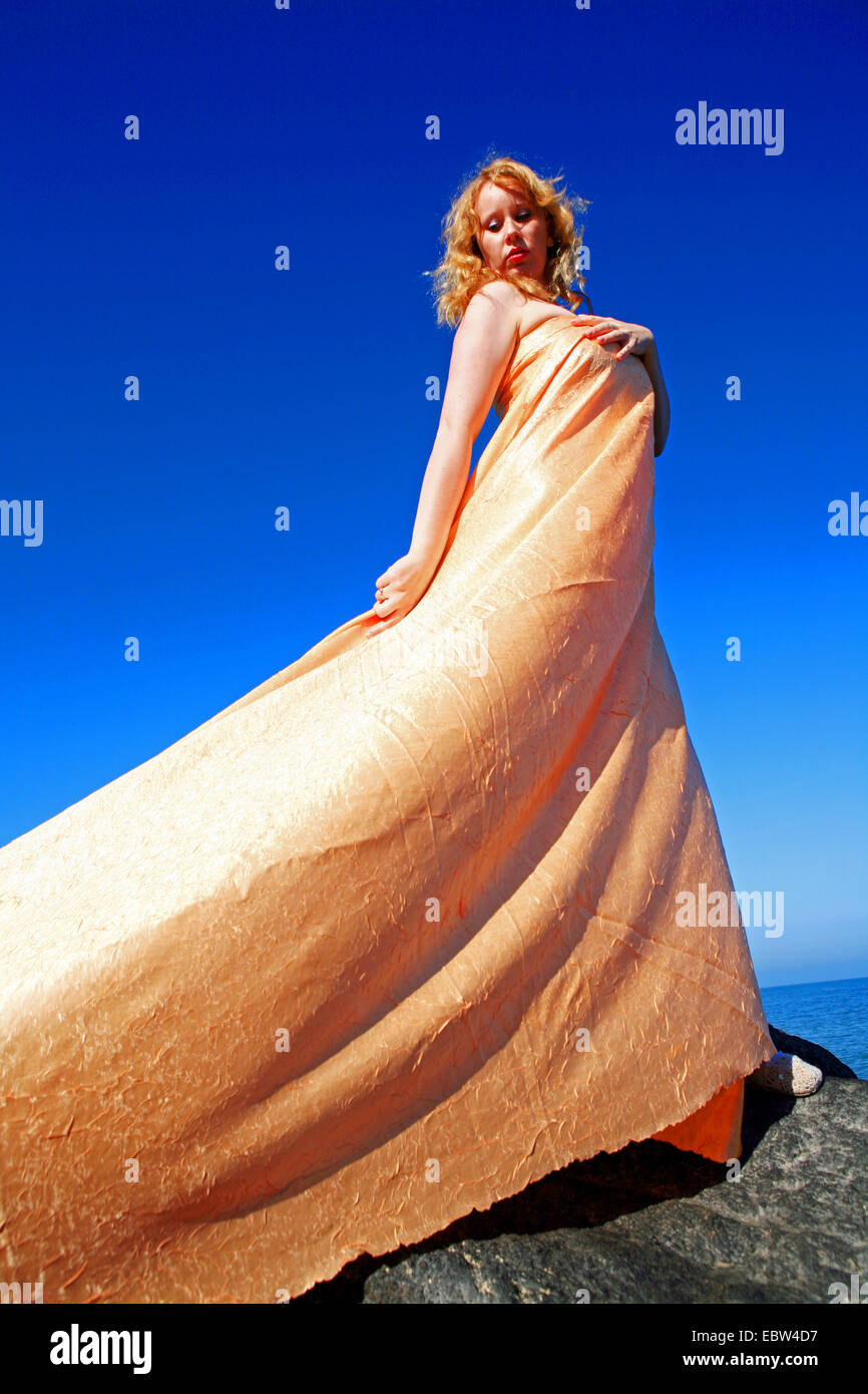 redheaded woman in a golden fabric standing on rock at the coast, Germany, Mecklenburg-Western Pomerania Stock Photo