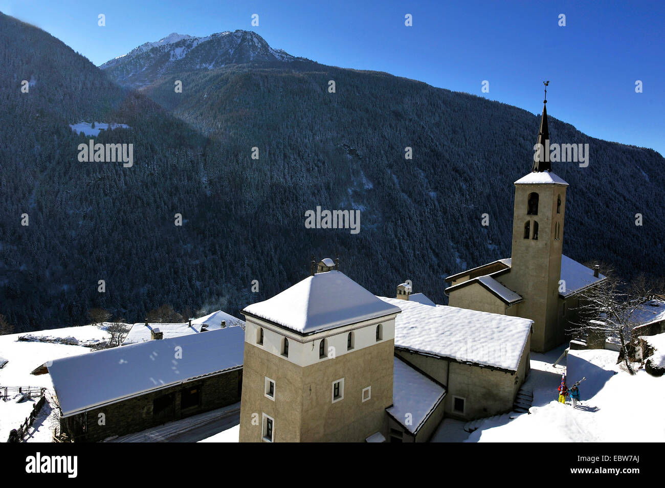 two wanderes with skiers passing church in front of impressive mountain sight, France, Savoie, Montvalezan Stock Photo