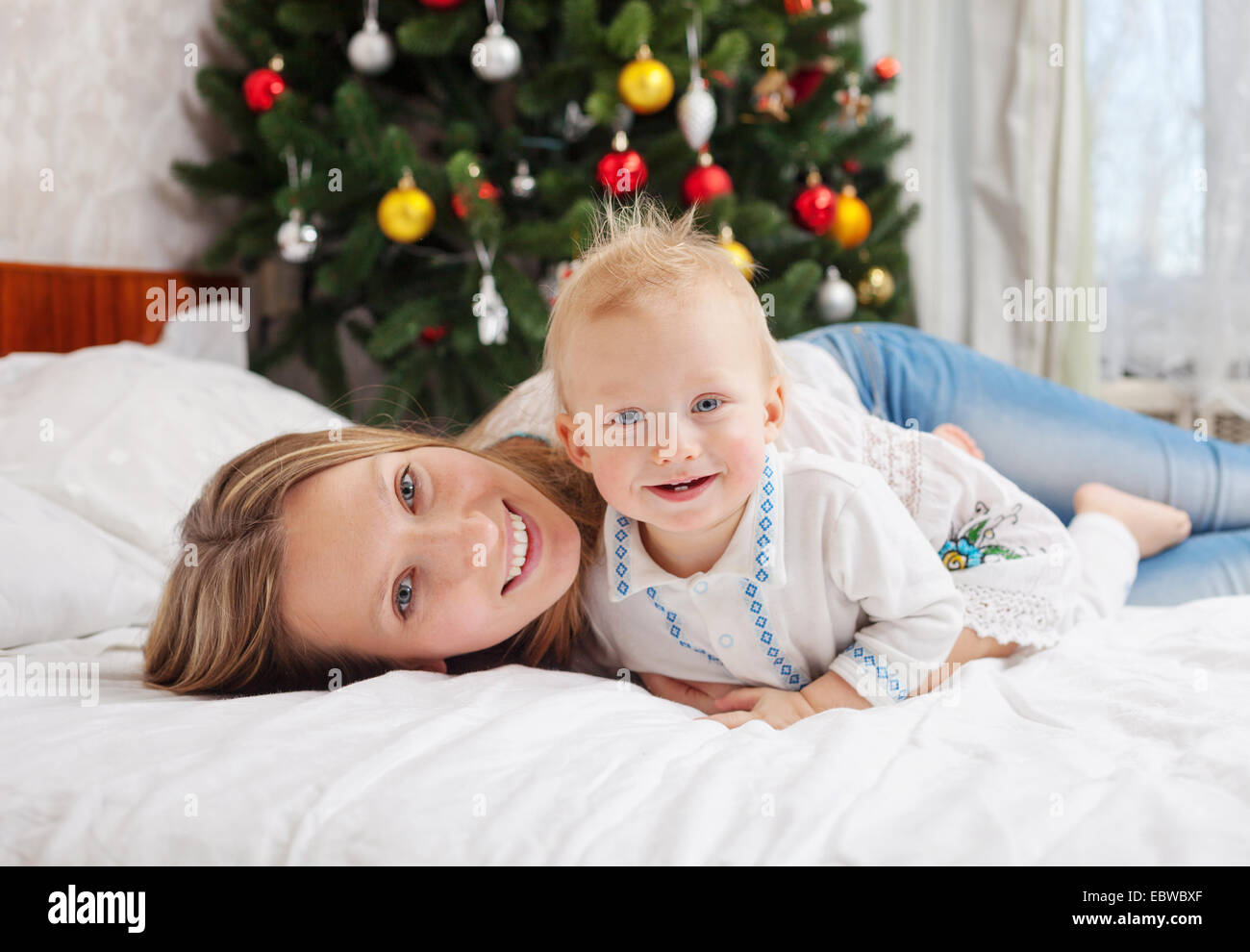 Portrait of happy mother and baby boy on bed at home with decorated Christmas tree in background Stock Photo