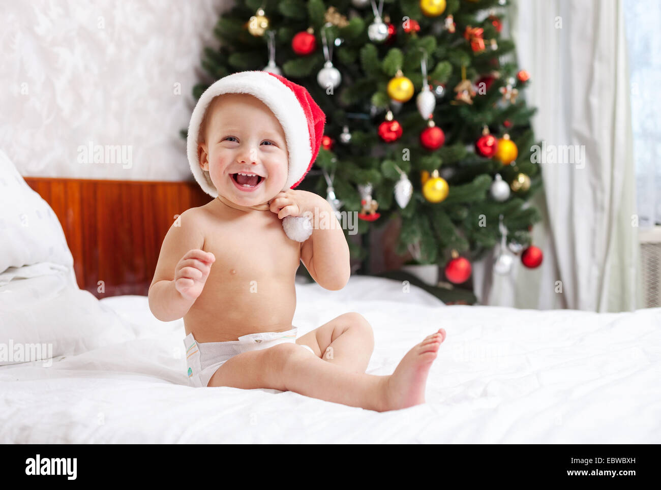 Christmas baby in santa hat sitting on bed and laughing Stock Photo