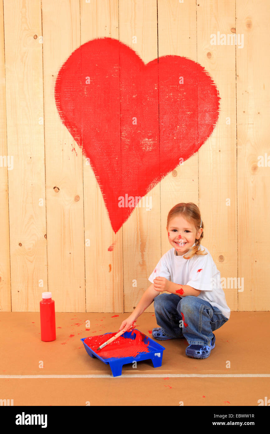 laughing little girl squatting in front of a wooden wall on which she has painted a heart with red paint Stock Photo