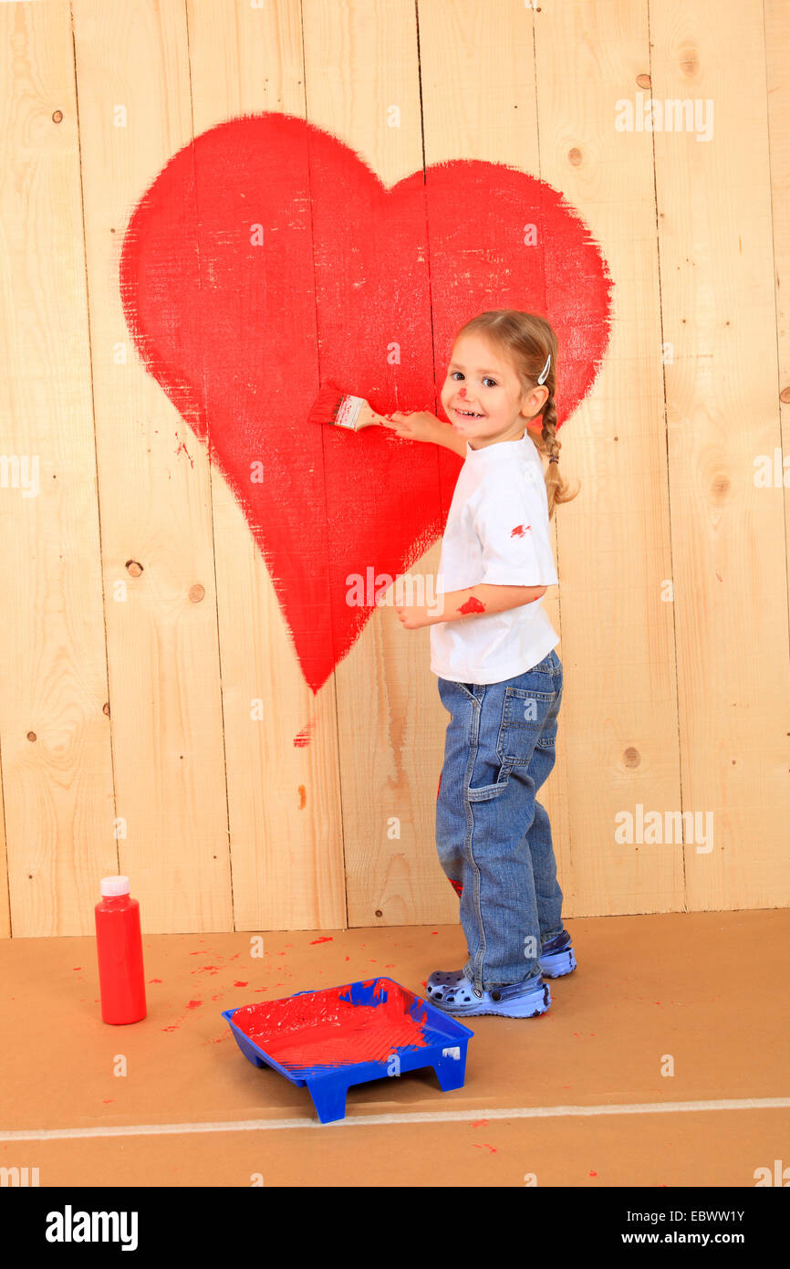 little girl painting a heart onto a wooden wall with red paint Stock Photo