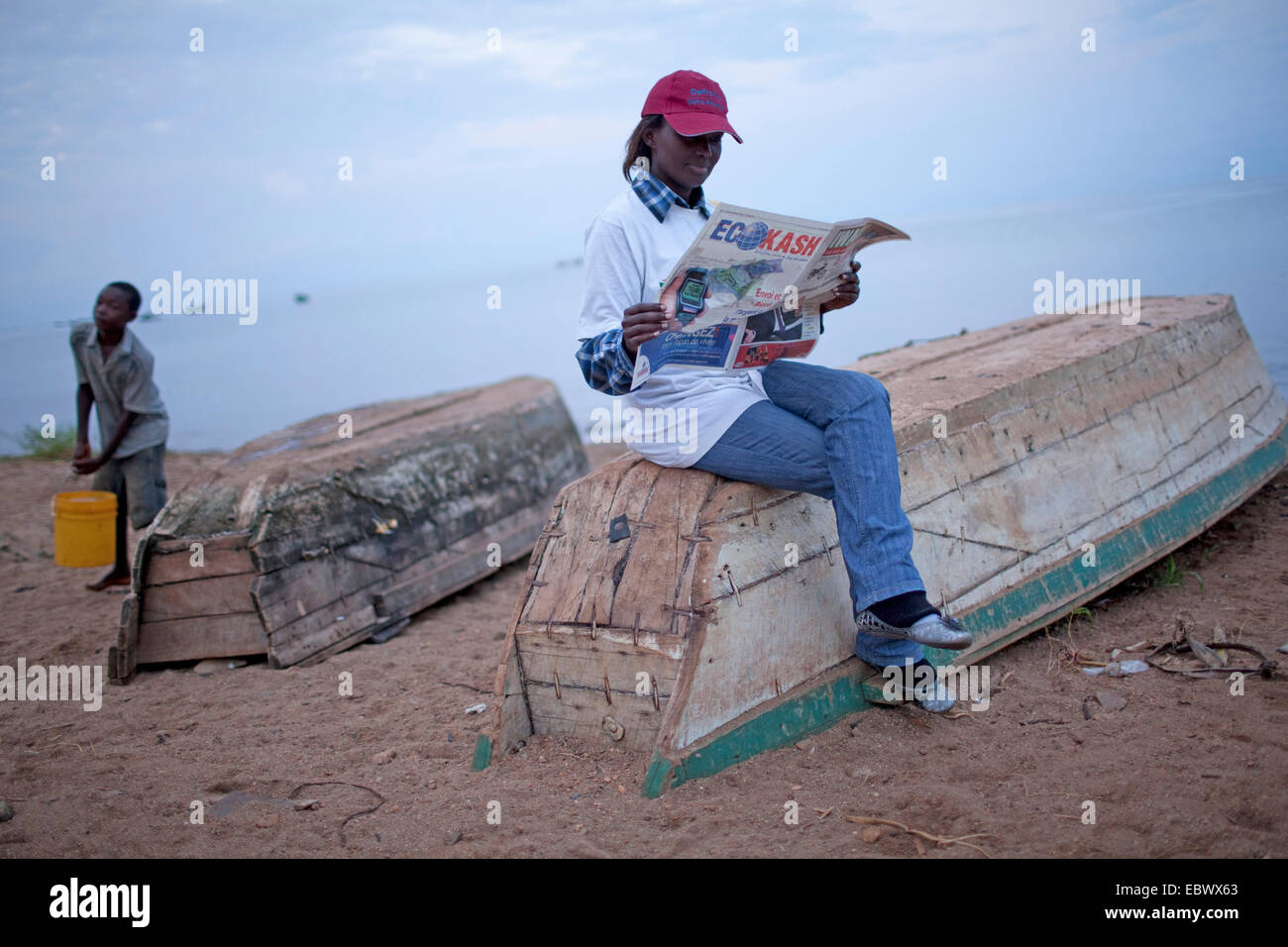 young woman sitting on an upside-down boat on the sand beach reading a paper while a boy is fetching water in a bucket from Lake Tanganijka, Burundi, Nyanza Lac, Mvugo, Nyanza Lac Stock Photo