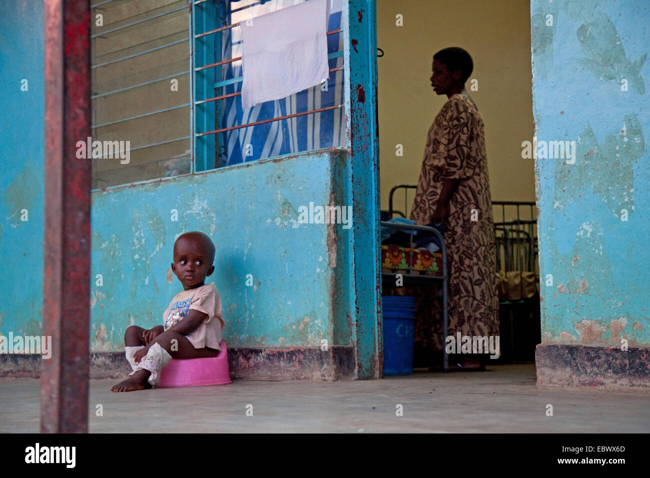 little boy sitting on a potty in front of an orphanage, woman inside is making beds, Burundi, Bujumbura Marie, Bujumbura Stock Photo
