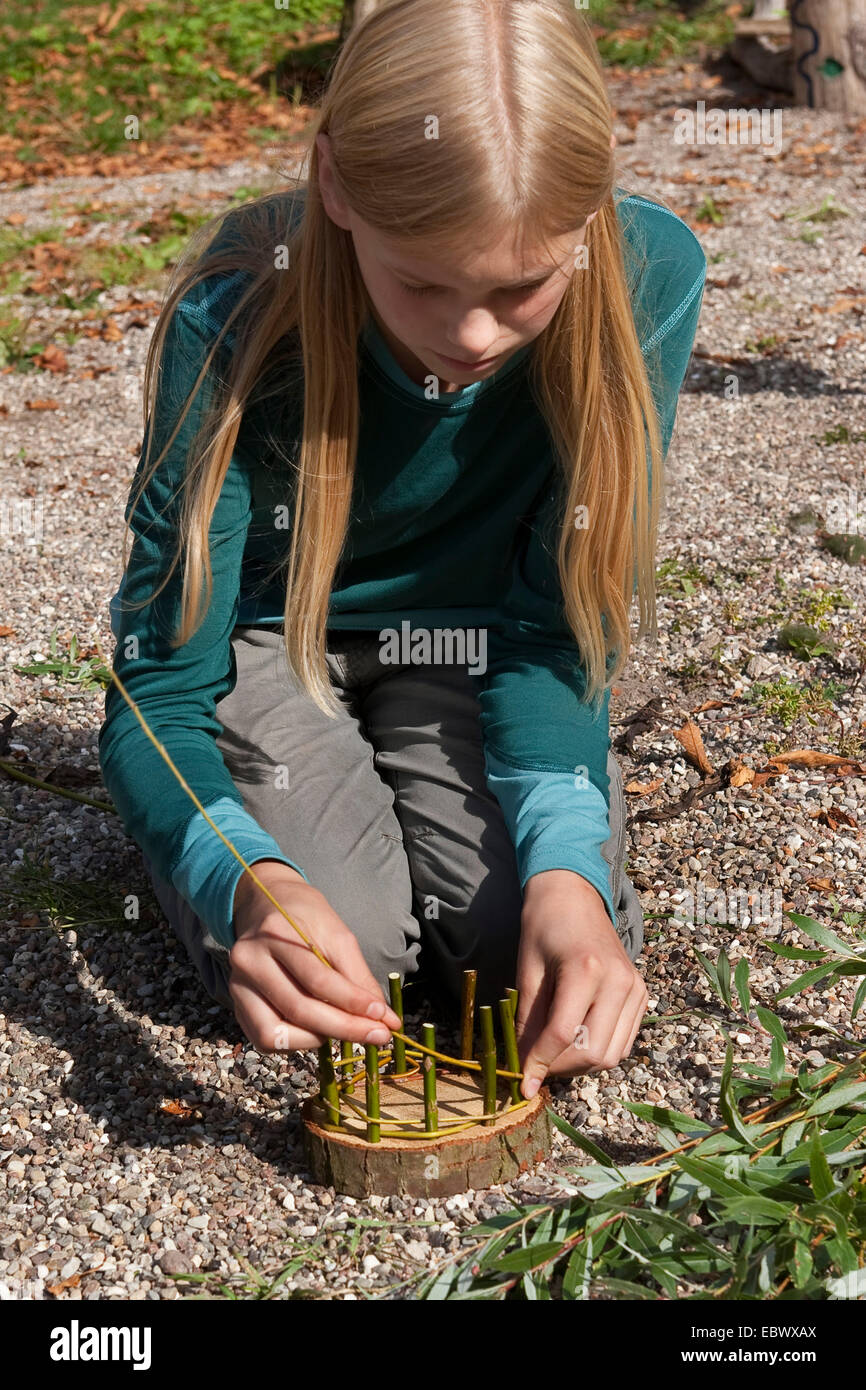 girl building an Easter basket from a tree disc, willow twigs, moss, daisies and coloured eggs; 3. step: weaving flexible willow twigs into the solide vertical ones, Germany Stock Photo