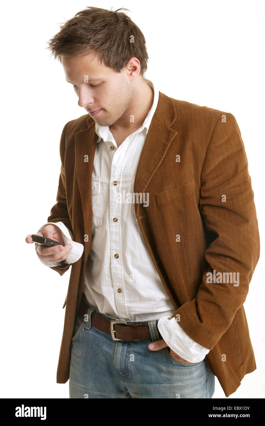 young man in a white shirt and brown jacket, writing an SMS with a hand in the pocket Stock Photo