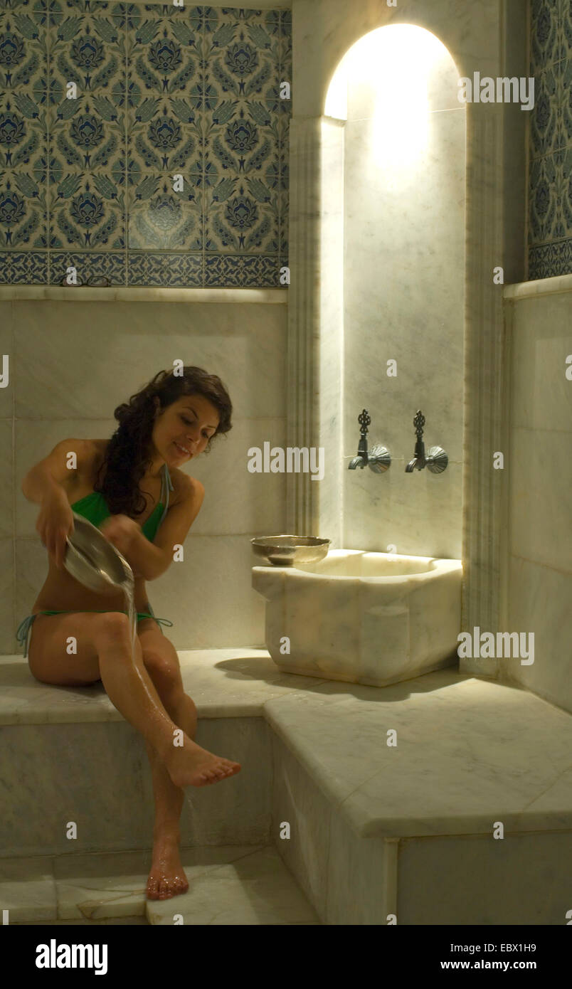 young woman sitting on a marmorn bench in a turkish bath pouring water on her legs from a metal bown Stock Photo