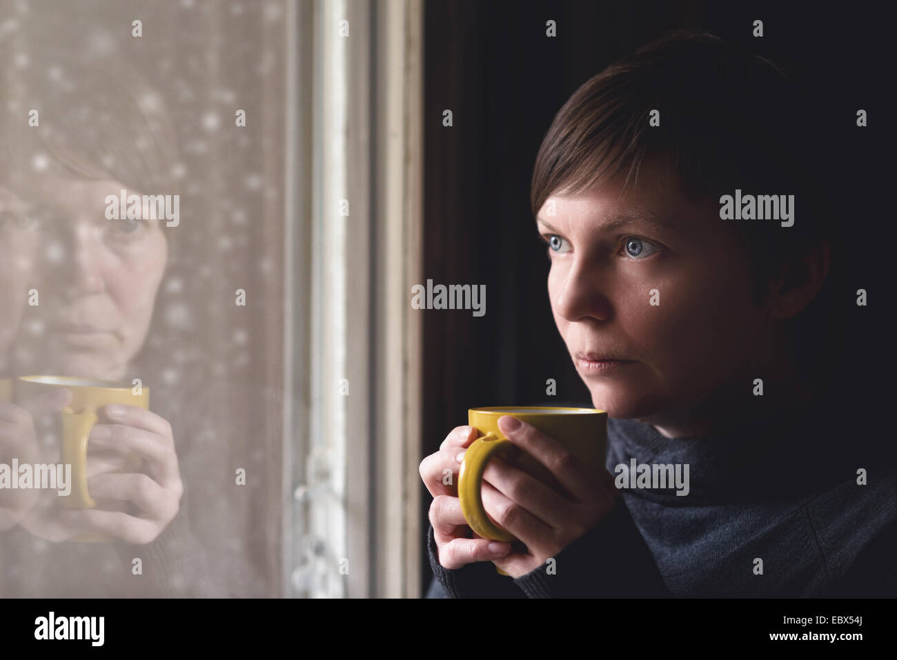 Lonesome woman drinking cup of coffee by the window of her living room, looking out at snow falling with a sad look on her face Stock Photo