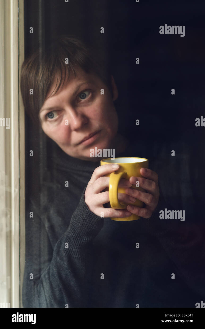 Lonely woman drinking cup of coffee by the window of her living room, looking out with a sad look on her face. Selective focus w Stock Photo