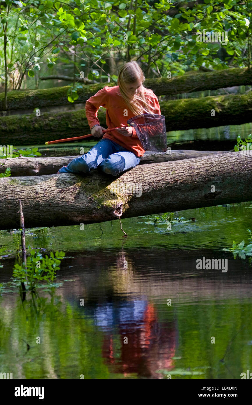 girl with dip net at a forest creek, sitting on a tree trunk over the water Stock Photo